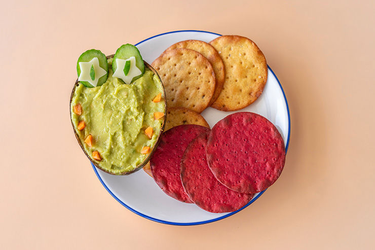 A plant-based meal with a Plate with guacamole decorated like a face, cucumber eyes, and colourful tortilla chips on a white plate over a peach background.