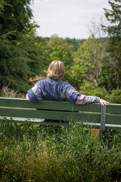 Auf dem Bild sieht man eine Person von hinten auf einer Bank sitzen und in die Weite auf einen Wald schauen.