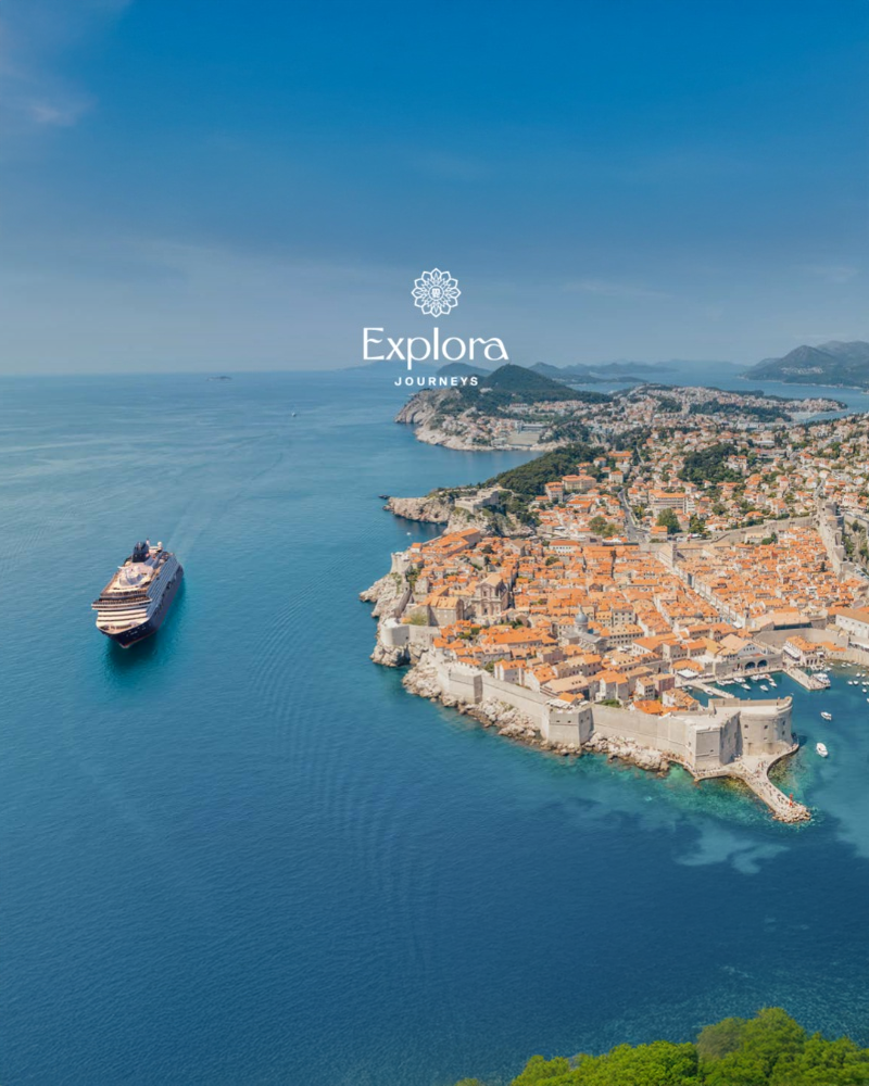 Cruise ship sails near a coastal city with orange rooftops and historic walls. Blue sea and sky. "Explora Journeys" text in the sky.