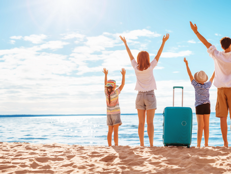 Family on sandy beach with raised arms, two suitcases nearby. Sunny day, blue sky and ocean in background. Joyful and carefree mood.