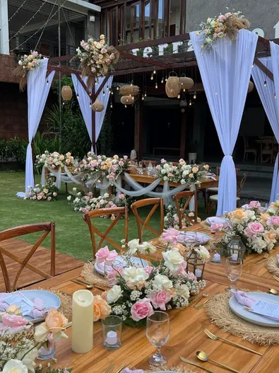 Outdoor wedding dining table setup on a lawn featuring pastel floral arrangements, place settings, and hanging lights.
