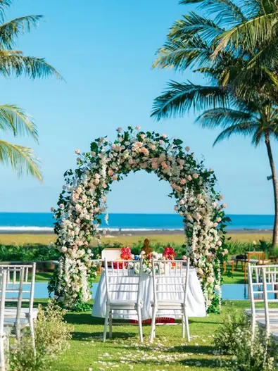Outdoor beachfront wedding ceremony setup with a floral arch, white chairs, and tropical palm trees overlooking the ocean.