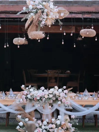 Wedding head table decorated with white drapes, pastel flowers, and rattan pendant lights.