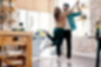 A joyful couple dances in a cozy kitchen with wooden shelves and a brick wall. Sunlight streams in, enhancing the warm, cheerful atmosphere.