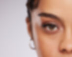 Close-up of a woman's eye with hoop earring, showcasing brown eye and full brows against a soft background, conveying a focused mood.