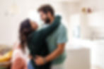A couple embraces in a brightly lit living room, smiling at each other. The room has a pink sofa, white cabinets, and a cozy atmosphere.