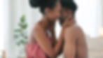 A couple smiling and embracing closely in a cozy room, she in a pink top. A plant and candle in the soft-lit background suggest warmth.