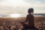 Person meditating on a sandy beach at sunset, with the reflective sea in the background creating a calm and serene atmosphere.