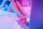 Blurred legs on a bed with pink lighting; a laptop and pink sex toys in the foreground. Relaxed and intimate mood.