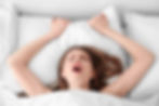 A woman lies in bed with her head on a white pillow, gripping it tightly, eyes closed and mouth open, expressing tension or stress.