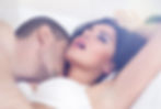 A couple is lying on a bed; one person kisses the other's neck. The woman appears relaxed, eyes closed, in a white bra against a soft background.