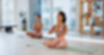 Two women practice yoga in a bright studio, sitting cross-legged in meditation pose. Shelves with candles and plants are visible. Peaceful mood.
