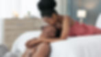 Woman in pink dress kisses man's forehead, both on bed in cozy room. Soft lighting, candles, and books create a warm, intimate mood.