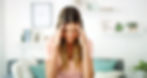 Woman with long hair holds head in hands, eyes closed in discomfort. She's in a bright room with shelves and plants. Wears striped top.
