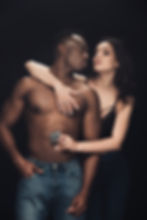 Man and woman in intimate pose; woman holds a condom near man's waist. Dark background, casual jeans, and black lingerie visible. Mysterious mood.