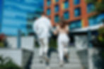 A man and woman holding hands climb outdoor steps, surrounded by greenery. Modern building with blue glass and brick facade in background.