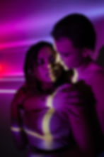 Two people, interracial couple embrace under purple and pink lighting, creating a romantic mood. Light patterns cover their faces and a netted top is visible.