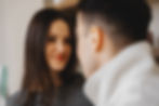 Woman smiling warmly at a man indoors. She wears a gray top, and he has a white jacket. Background is softly blurred, creating a cozy mood.