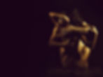 Two nude individuals embrace intimately in a dimly lit setting, their bodies intertwined in a sensual and artistic pose. The golden-toned lighting highlights the curves of their muscles and skin, creating a dramatic and passionate atmosphere against a dark background.