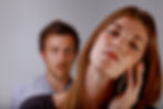 Woman with long hair on phone, looking indifferent, as a man with soft focus stands behind her. Neutral background, serious mood.