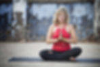 Woman in a red top meditating in a seated position on a mat outdoors, with a calm expression. Blue textured wall in the background.