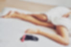 Person lying under a white duvet on a bed, showing legs. Nearby are black lingerie and a pink item, suggesting a relaxed intimate setting.