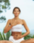 Woman meditating outdoors with eyes closed, one hand on chest and one on abdomen. Wears white top and shorts. Peaceful, green background.