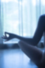 Silhouette of a person meditating indoors, seated with fingers in a mudra. Soft light filters through sheer curtains, creating a calm mood.