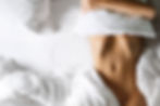 A person lying on a bed covers their face with a pillow, surrounded by white sheets. The mood is relaxed and calm.