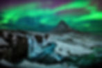 Snow-covered mountain landscape in Iceland with a waterfall in the foreground, illuminated by the vibrant green and purple glow of the aurora borealis under a starry night sky.