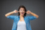 Woman in denim shirt smiles playfully, plugging ears with fingers against gray background. Casual and cheerful mood.