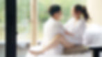 Abstract view of a couple in white robes sitting closely on a bed, blurred background with greenery outside a window, soft and intimate mood.