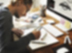 Man drawing at a wooden desk with markers and a notebook. A computer displaying a "LIFESTYLE" graphic is in the background. Creative mood.