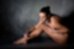 A woman applies intricate body art to her leg in a dimly lit room with a textured gray wall. She appears focused and calm.