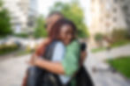 Two people hugging warmly on a city street, one smiling. Green trees and buildings in the background. A joyful, friendly moment.