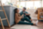 Couple sitting on kitchen floor, smiling and toasting with red wine. Bright kitchen with blue cabinets, wooden shelves, and plants. Cozy mood.