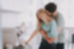 Couple embracing in a bright kitchen, smiling near a coffee maker on a counter. Both wear light blue-green shirts, creating a warm, cozy mood.