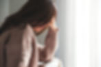 A woman in a beige sweater sits with her head in her hand, appearing contemplative. Soft light filters through white curtains in the background.