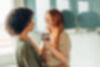 Two people in a kitchen share a moment. One with curly hair, the other with red hair, holding wine glasses, smiling warmly at each other.