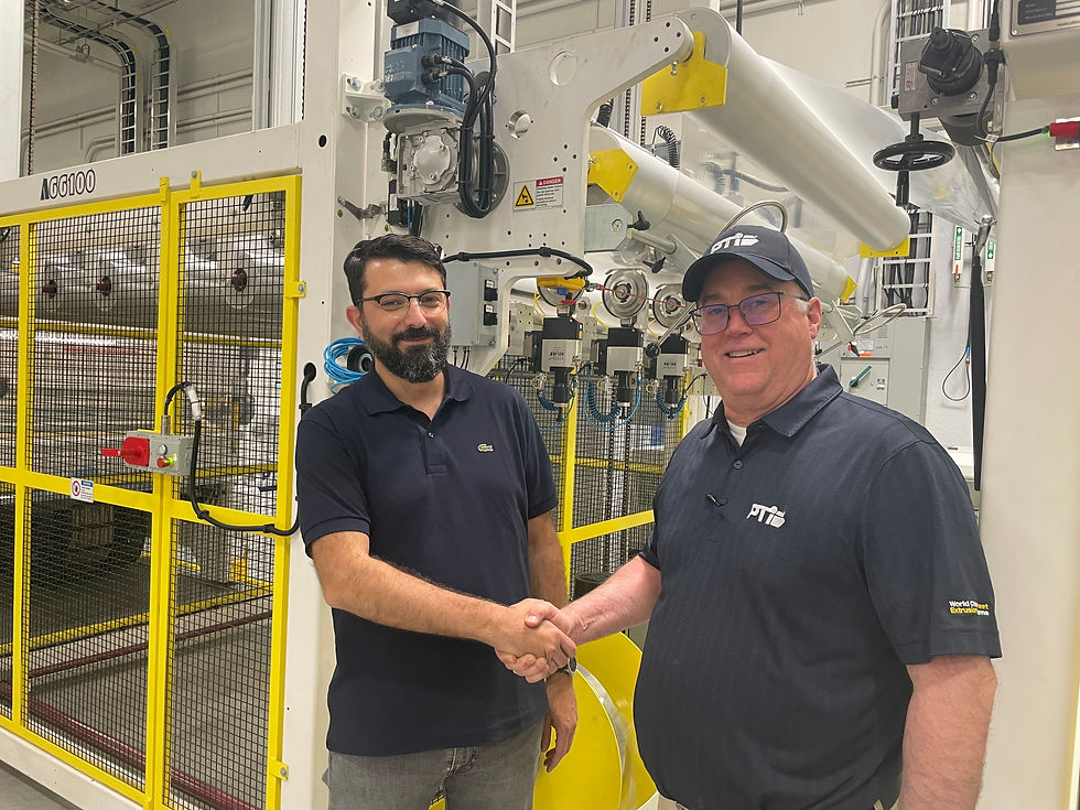 Two men in navy polos shake hands and smile beside a large yellow-and-white industrial machine in a factory.