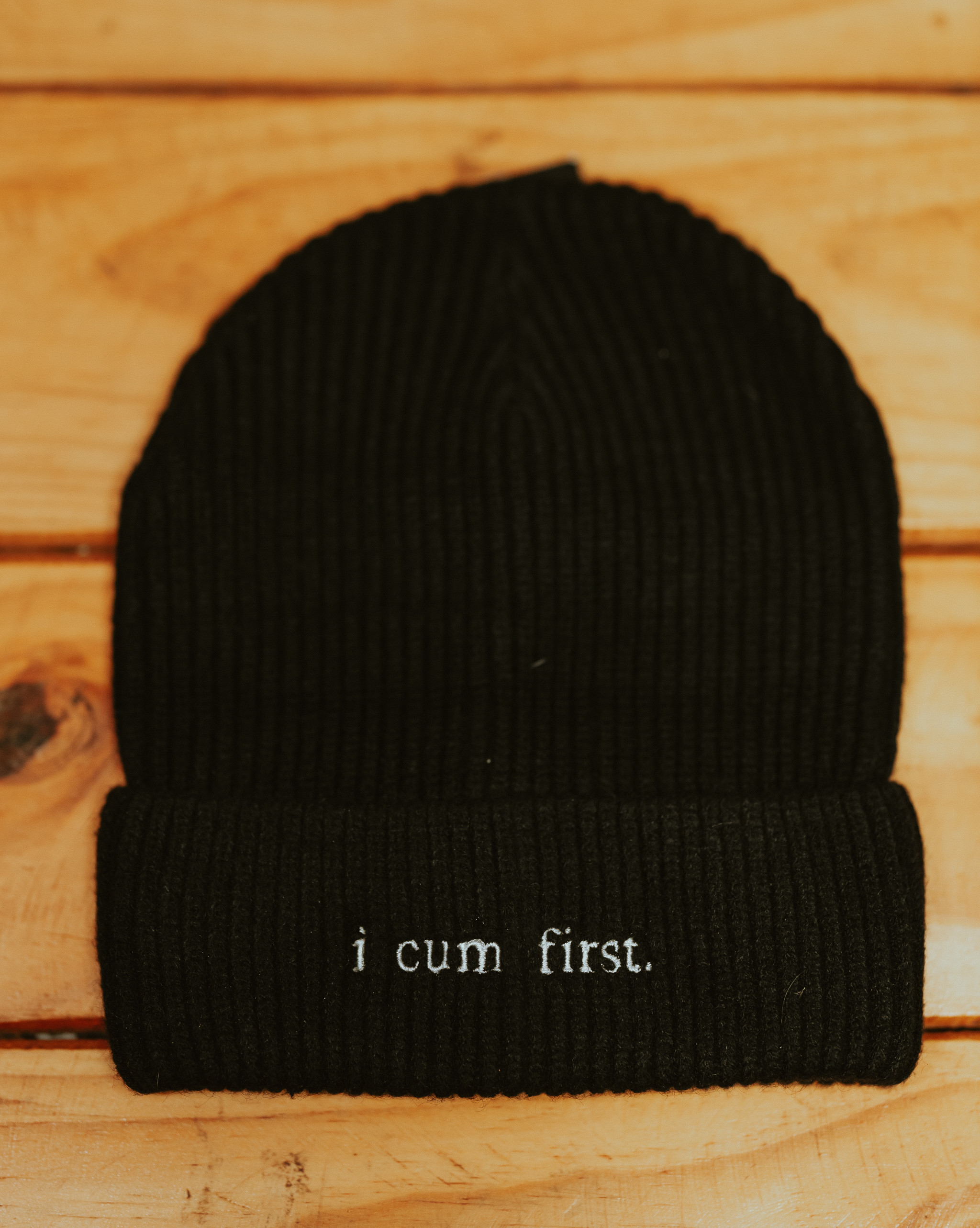The I CUM FIRST beanie on a wooden background