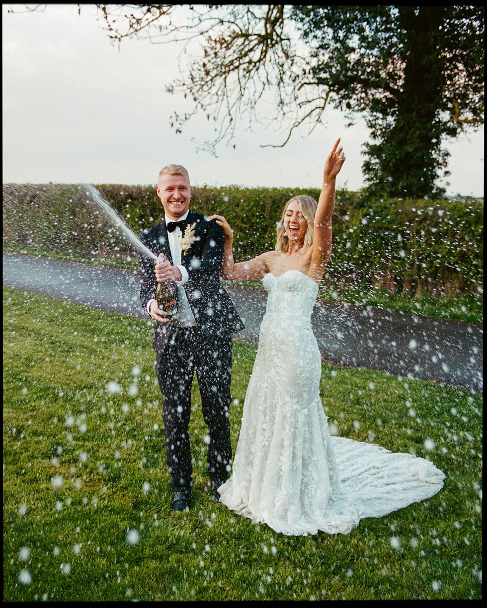 Caught in a jubilant burst of champagne spray, a newlywed couple celebrates their union with beaming smiles. The groom, in a smart black suit, is popping a bottle, while the bride, in a strapless lace gown, throws her arms up in elation. The spontaneous moment is framed by the verdant backdrop of a neatly trimmed hedge and a sprawling lawn, with the effervescent droplets catching the light, creating a shower of sparkling detail that gives life to the photo, much like the vivid colors and textures often brought out by Portra 400 film. Their expressions of pure joy are the centerpiece of this lively and memorable celebration.