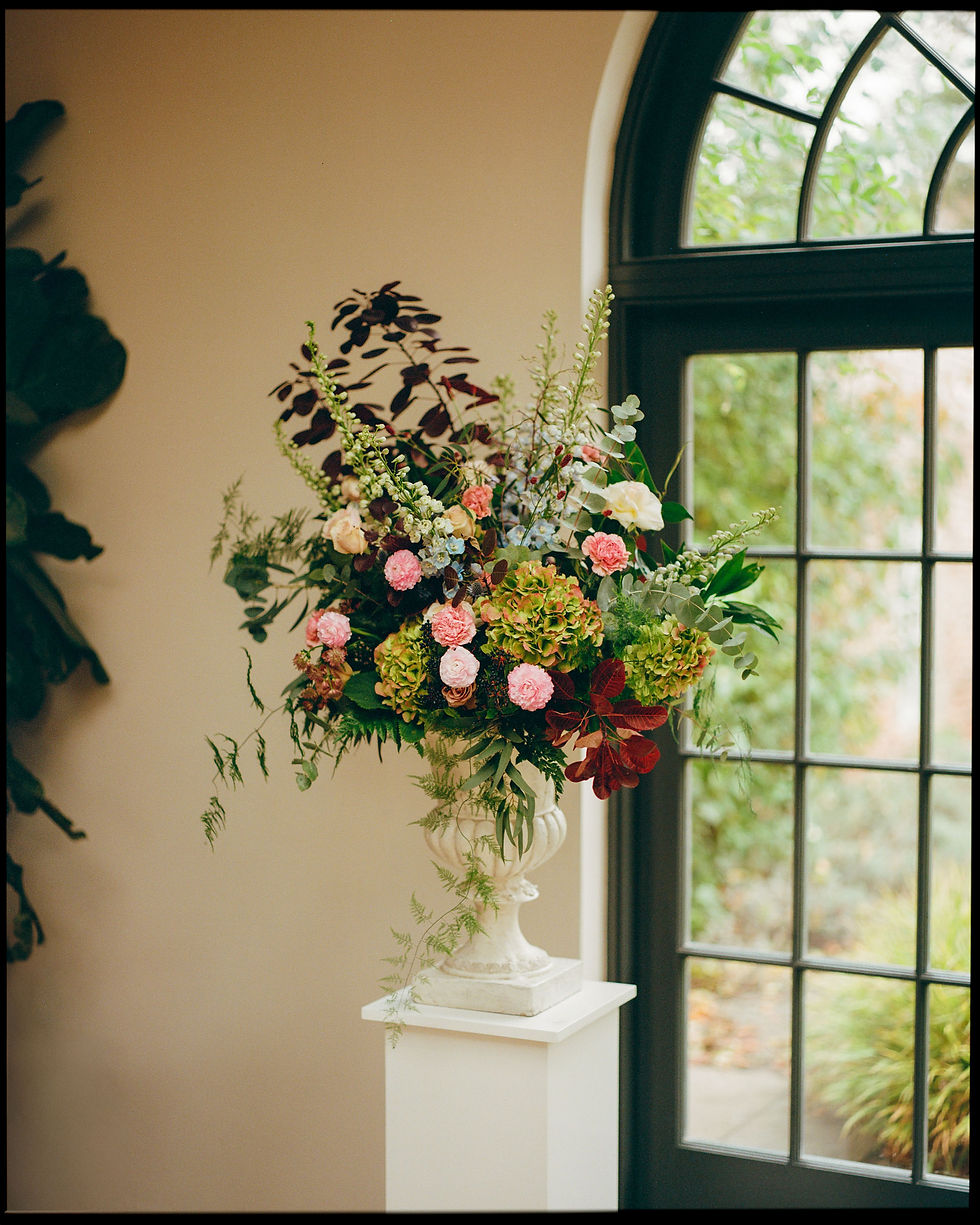 A richly detailed floral display graces a stately white pedestal vase, positioned beside a window that frames the verdant tranquility outside. The arrangement is a harmony of color with pink roses, deep red flowers, and varied greenery, all captured in the soft, warm tones characteristic of a Portra 400 film photograph. The gentle light from the window bathes the scene, accentuating the textures and hues of the petals and leaves, contributing to the arrangement's natural elegance. This image, with its inviting composition, conveys a timeless beauty and a sense of peacefulness.