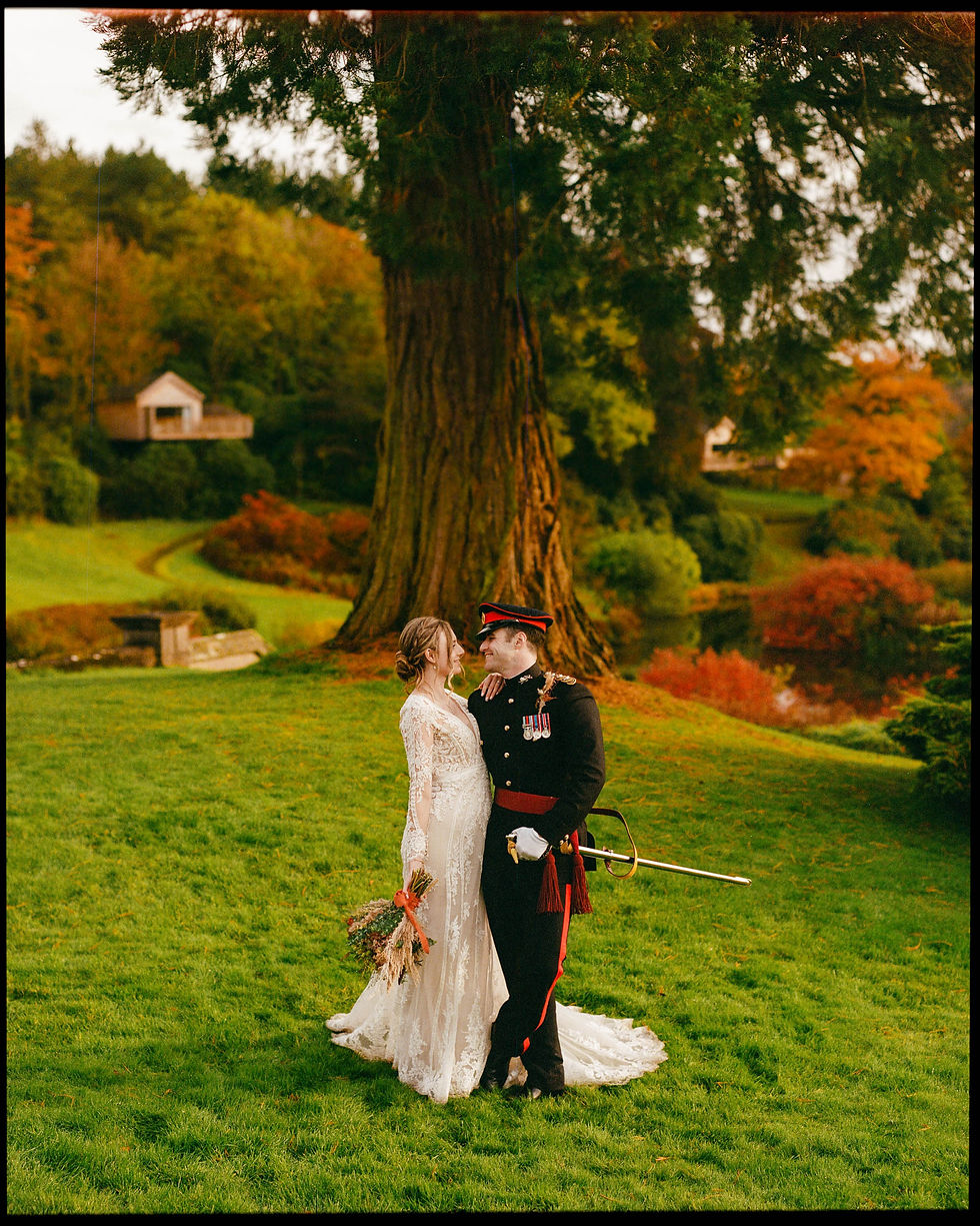 In this portrait, suffused with the rich, warm palette of Portra 400, a bride and groom share an intimate gaze, amidst a lush, autumnal landscape. The bride, in a lace gown, holds a bouquet that mirrors the earthy tones around them. The groom stands distinguished in a ceremonial military uniform, complete with red accents and a sword at his side, which adds a sense of tradition and formality to the scene. The majestic tree under which they stand anchors the composition, symbolizing strength and endurance, much like the commitment they have just made to each other. The overcast sky casts a soft, diffused light, perfect for capturing this moment of tender connection.