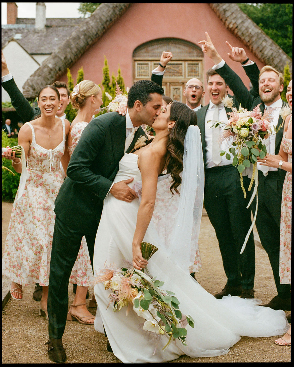 Captured in the vibrant and warm hues that evoke the timeless essence of Portra 400 film, a bride and groom share a kiss, encapsulating the joy of their wedding day. The bride, clad in an elegant white gown with a long veil, holds a whimsical bouquet, complemented by the groom's classic black suit and tie. Around them, a lively group of wedding guests celebrates the moment; their elated expressions and dynamic poses add to the festive atmosphere. The scene is set against a quaint architectural backdrop that lends a touch of historical charm to this snapshot of love and celebration.