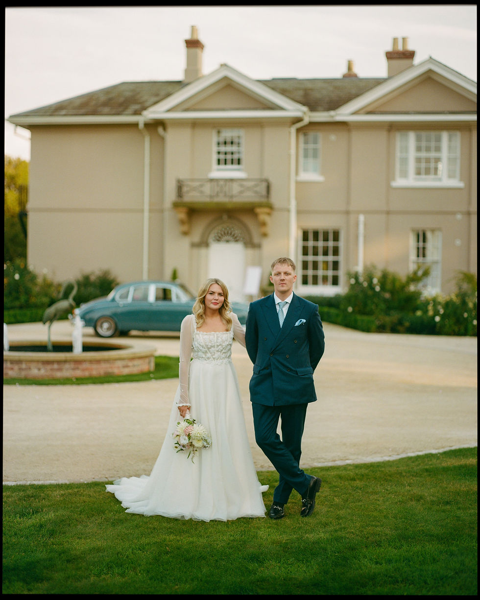 A bride and groom stride confidently across a manicured lawn, with the bride holding a delicate bouquet, complementing her graceful white gown. The groom is sharp in a teal suit, imbued with the depth of color that Portra 400 film brings to the green hues of his attire. Behind them, an elegant estate provides a stately backdrop, its symmetry and classic design adding to the formality of the occasion. The overcast sky softens the light, allowing the couple's poised expressions and the romantic details of their wedding attire to take center stage in this timeless photographic moment.