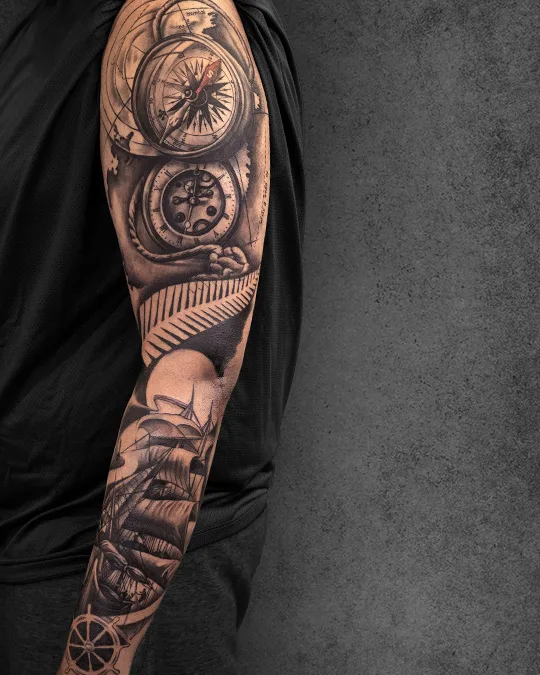 Realistic full-sleeve tattoo on a man’s arm, showcasing a compass, maps, and a ship, embodying exploration and nautical travel.