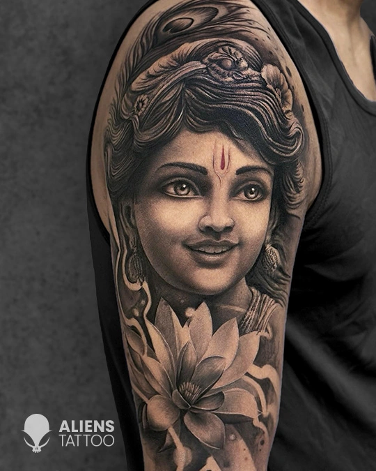 Realistic black and grey goddess portrait tattoo with soft facial details and lotus flower design on upper arm, crafted by Aliens Tattoo, the best tattoo studio in pune.
