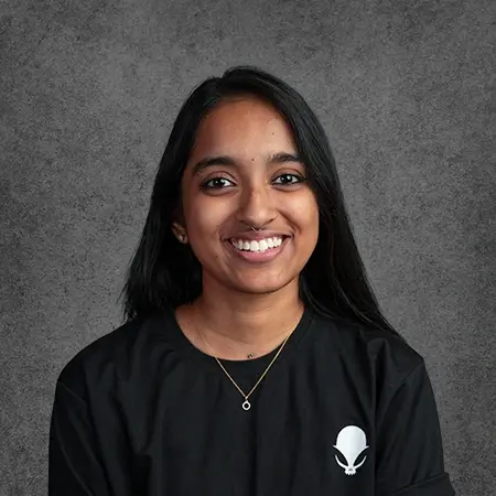 A professional portrait of Junekha Mary Jacob, an expert tattoo artist at Aliens Tattoo, the best tattoo studio in Kochi, smiling in a branded black t-shirt against a textured grey studio backdrop.