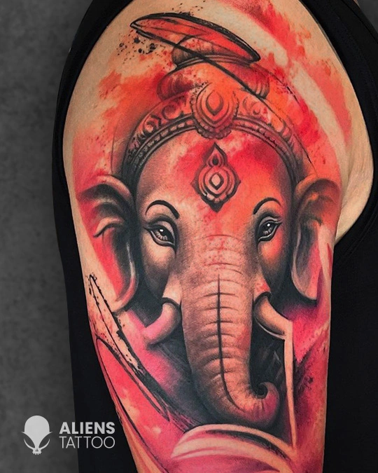 Hyper realistic Lord Ganesha tattoo with bold red watercolor effects on upper arm, showcasing detailed Hindu deity artwork by Aliens Tattoo, the best tattoo studio in pune.